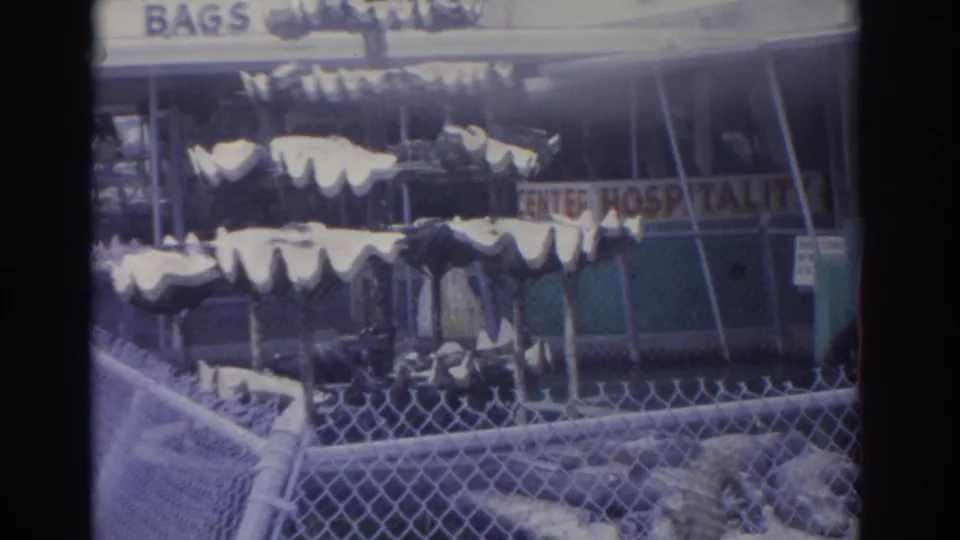 1962 Florida home-movie frame showing pale alligator bags outside a shop, with chain-link fencing and partial sale signage visible.