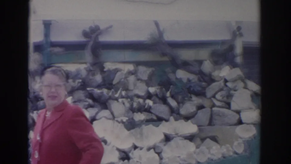 The woman remains near the stone wall, facing the camera in soft, faded 8mm color from the Florida clip.