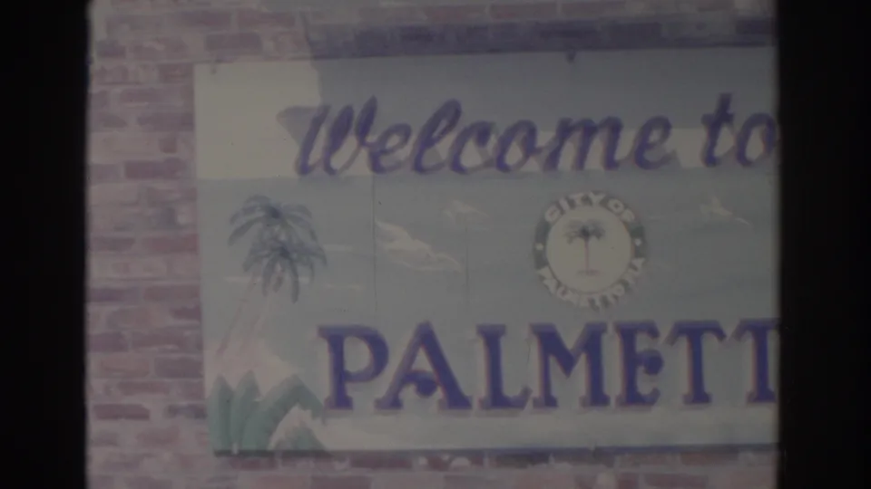A painted Welcome to Palmetto sign with palm graphics appears beside brickwork in the 1962 Florida home-movie frame.