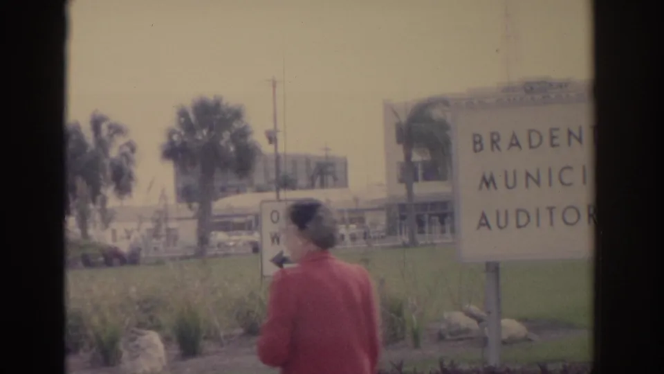 1962 Florida home-movie frame with a woman in red walking past the Bradenton Municipal Auditorium sign and palm-lined grounds.