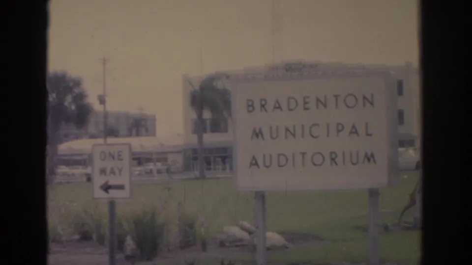 The Bradenton Municipal Auditorium sign stands on landscaped grounds with a one-way street sign and mid-century building behind it.