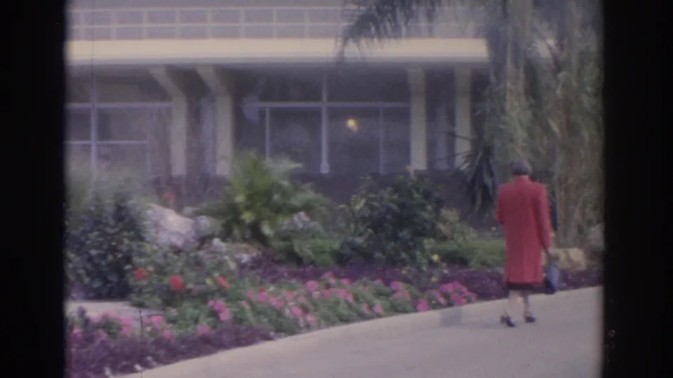 1962 Florida 8mm frame showing a woman in a red coat walking through manicured garden beds near palms and a building.