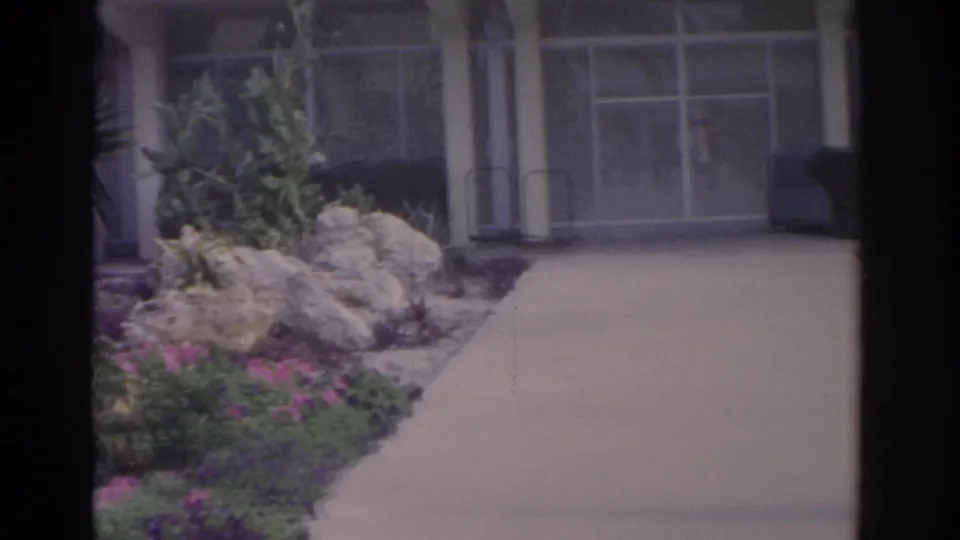 1962 Florida home-movie frame with a broad walkway, landscaped rocks, tropical planting, windows, and a shaded entrance area.