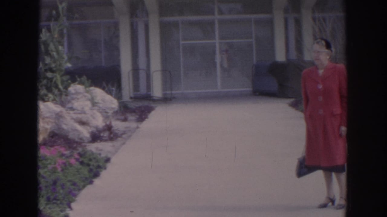 Woman in a red coat stands on a garden walkway near flowers and a glass-fronted building.