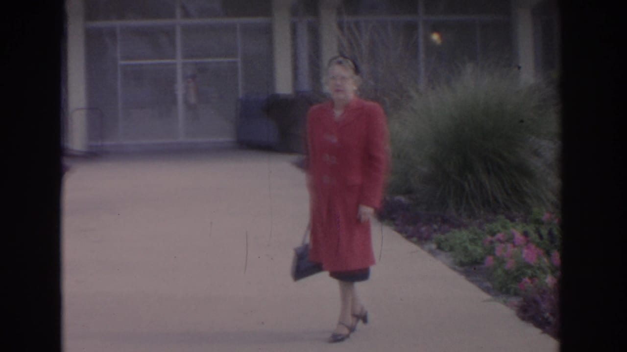 Woman in a red coat walks along a paved path beside shrubs and flowers.
