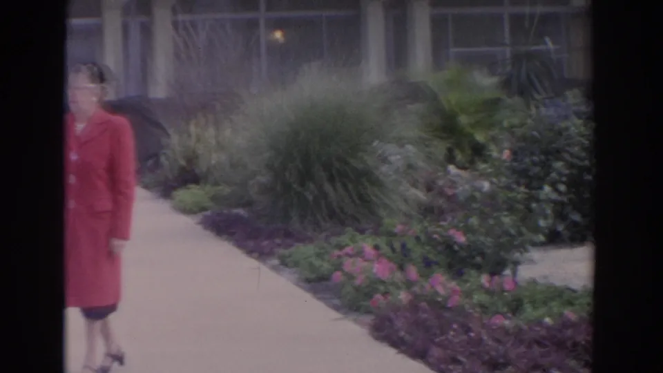 A partially framed woman in a red coat passes the camera along a landscaped path in 1962 Florida home-movie footage.