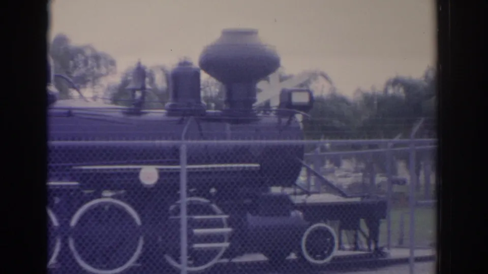 Black steam train replica in a Florida park, filmed from outside a chain-link fence on 8mm color film.