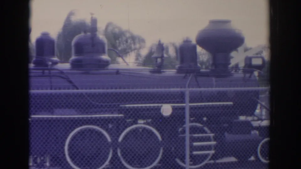 8mm frame of a black train replica display, with roof fixtures, large wheels, and fence lines visible in Florida park surroundings.
