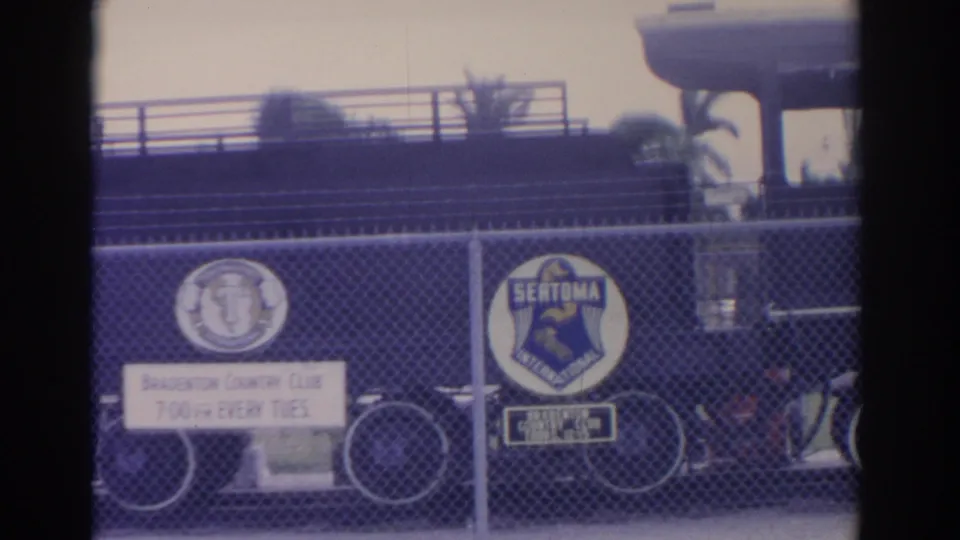 1962 Florida 8mm frame with Bradenton Country Club signage and a Sertoma International emblem on fencing before a dark locomotive.