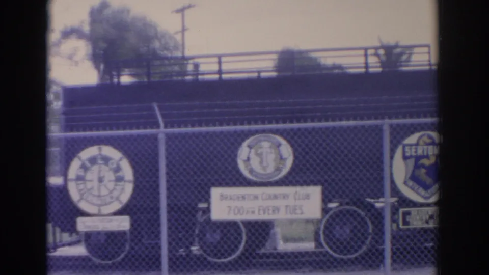 1962 Florida 8mm frame of exterior club fence signage, including Bradenton Country Club text and a Sertoma emblem near a bicycle.