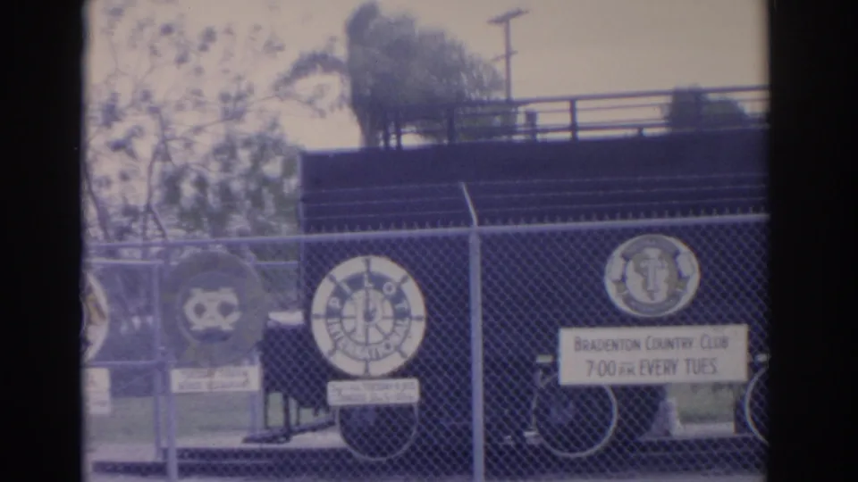 1962 Florida 8mm frame showing several club emblems on a chain-link fence, with Bradenton Country Club signage and a bicycle behind it.