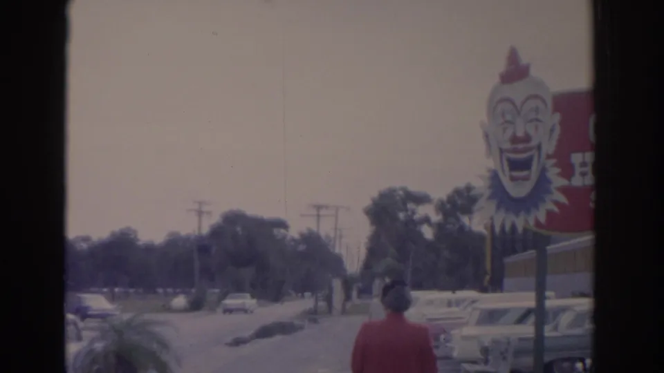 A woman in a red jacket walks along a Florida street near a large clown-faced circus billboard, with parked cars and trees nearby.