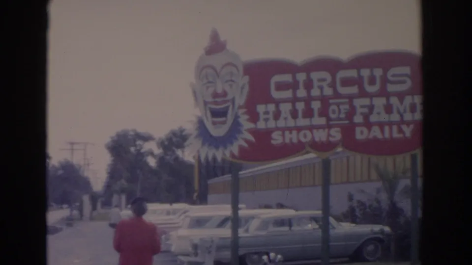 A large clown-faced Circus Hall of Fame sign stands over parked cars while the woman in red remains in the foreground.