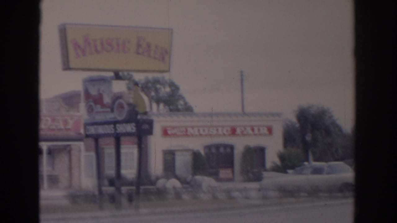1962 Florida home-movie frame showing the Music Fair sign, classic car display graphic, and a parked car near the attraction exterior.