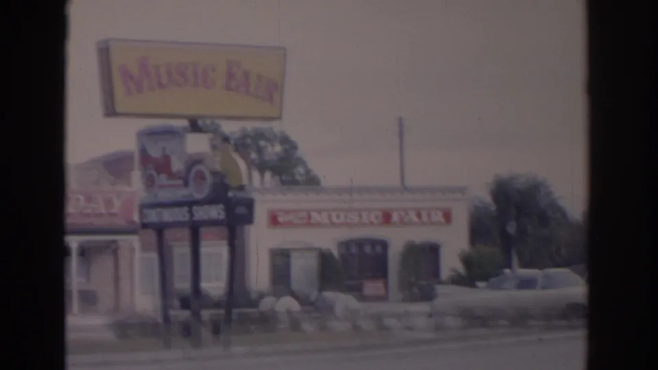 1962 Florida home-movie frame showing the Music Fair sign, classic car display graphic, and a parked car near the attraction exterior.