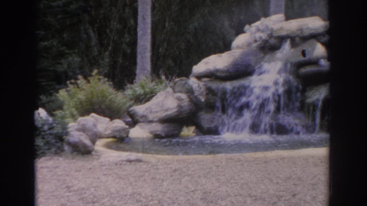 1962 Florida 8mm frame showing a rock waterfall cascading into a small pond near gravel and garden shrubs.