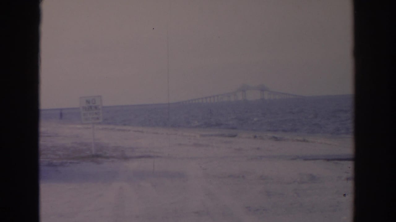 1962 Florida coastal home-movie view with a No Parking sign, pale sandy ground, choppy water, and bridge span on the horizon.
