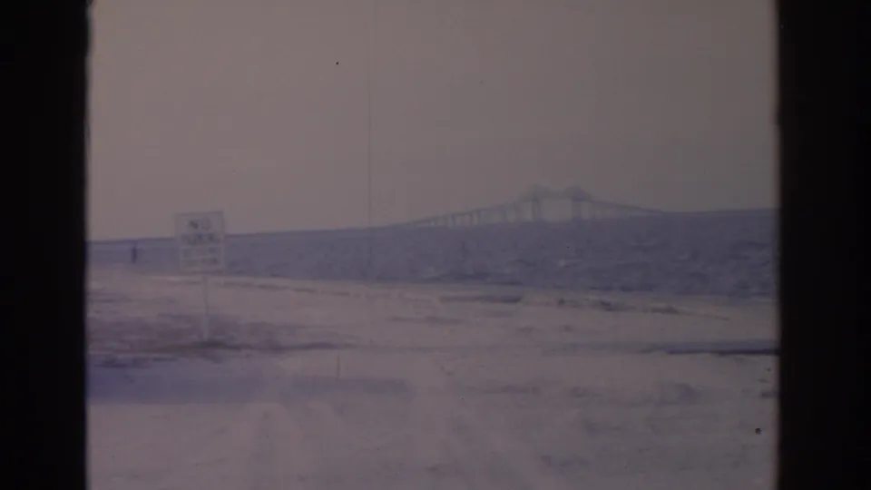 1962 Florida coastal home-movie view with a No Parking sign, pale sandy ground, choppy water, and bridge span on the horizon.