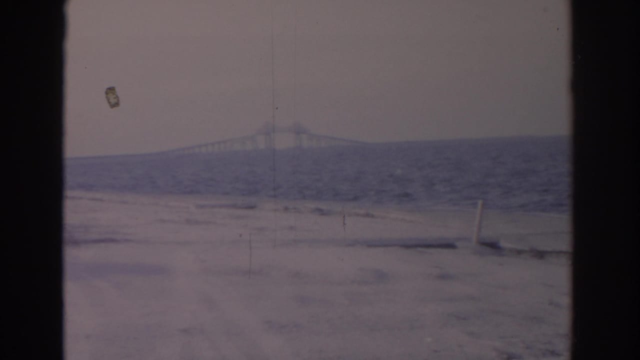 Faded 1962 Florida 8mm frame looking across a sandy coastal edge toward rough water and a long bridge in the distance.