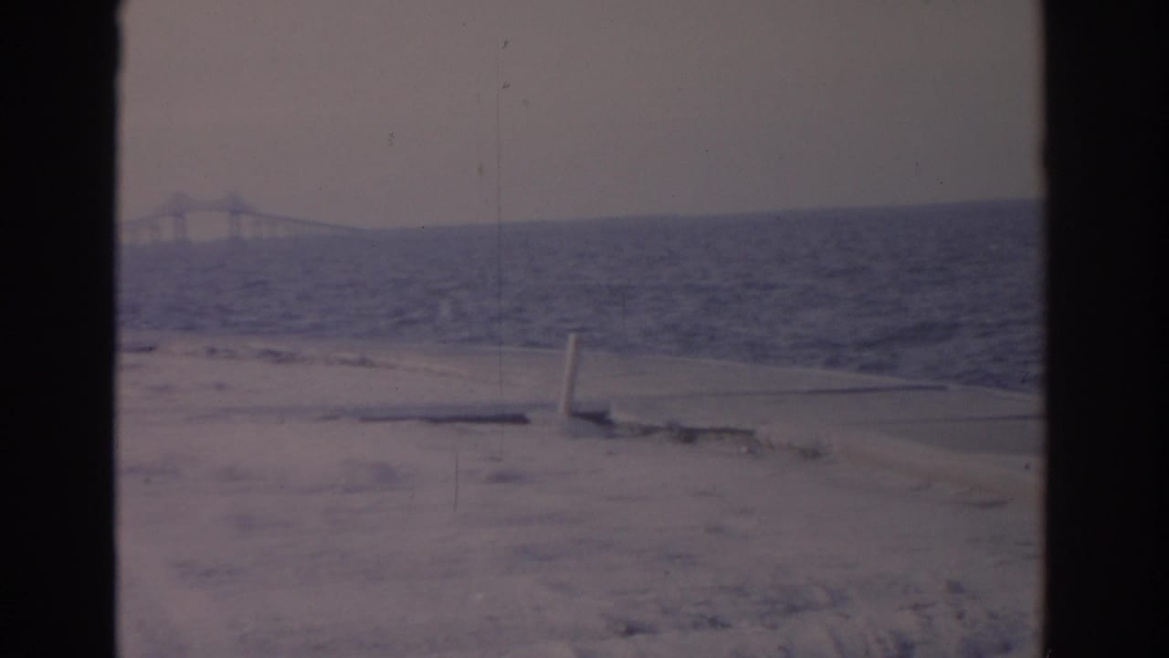 1962 Florida home-movie frame showing a sandy foreground, choppy coastal water, and a bridge span faintly visible on the horizon.
