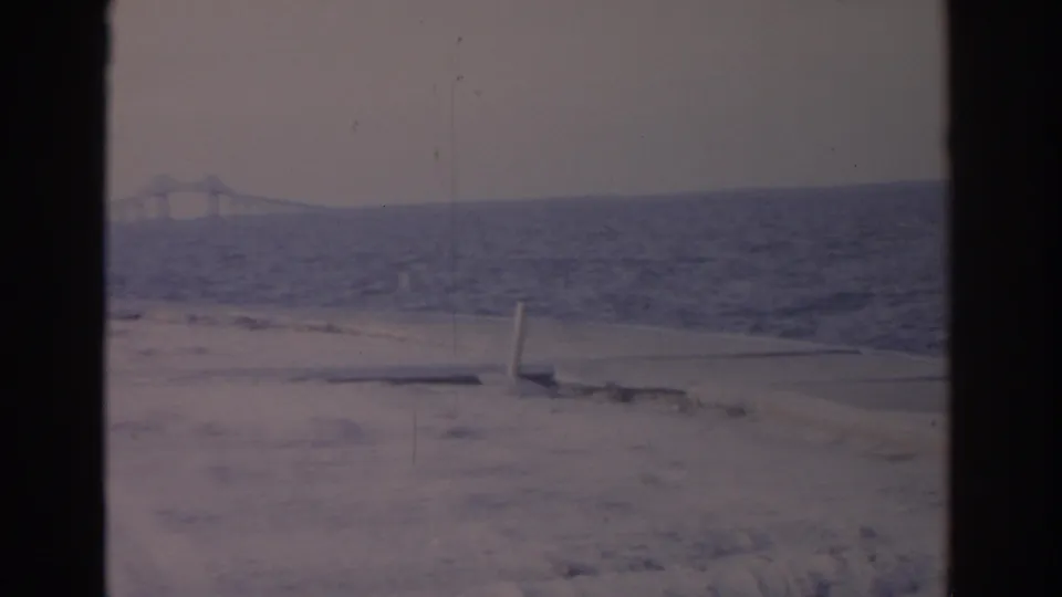 1962 Florida home-movie frame showing a sandy foreground, choppy coastal water, and a bridge span faintly visible on the horizon.