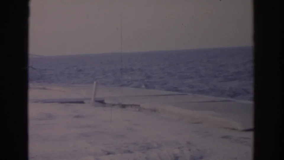 1962 Florida 8mm frame showing snowy sand, ocean water, pale sky, and a low concrete edge near the shoreline.