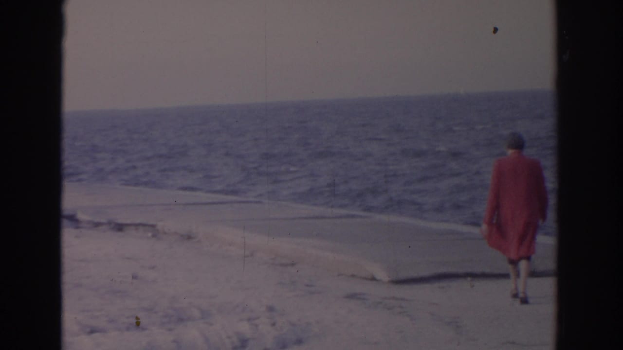 1962 Florida home-movie frame of an adult woman in a red coat walking beside snowy sand with the ocean behind her.