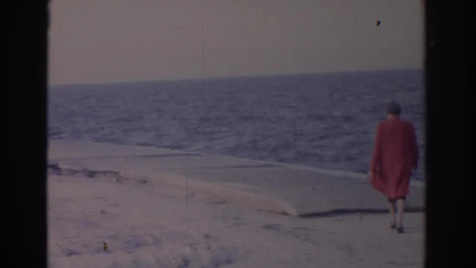 1962 Florida home-movie frame of an adult woman in a red coat walking beside snowy sand with the ocean behind her.
