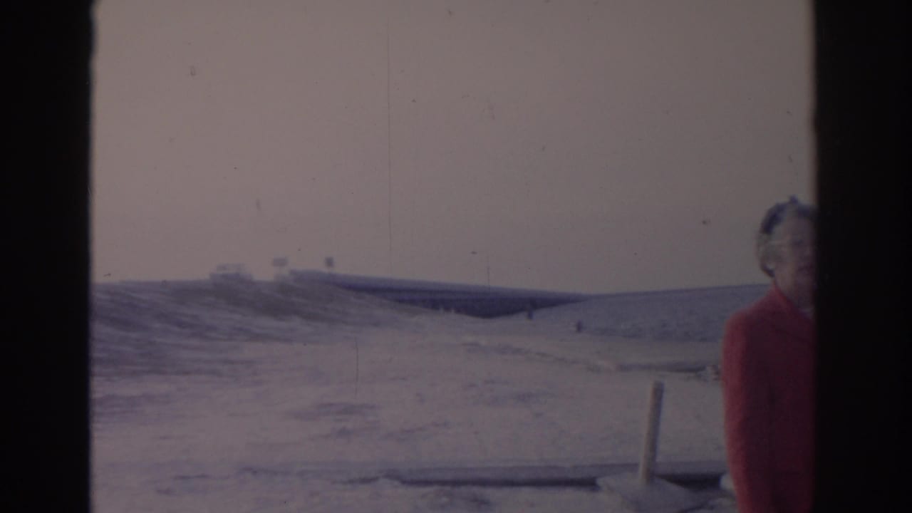 1962 Florida 8mm frame showing a red-coated adult woman near the camera, snowy beach, and a distant bridge over water.