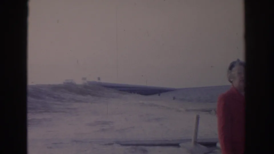 1962 Florida 8mm frame showing a red-coated adult woman near the camera, snowy beach, and a distant bridge over water.