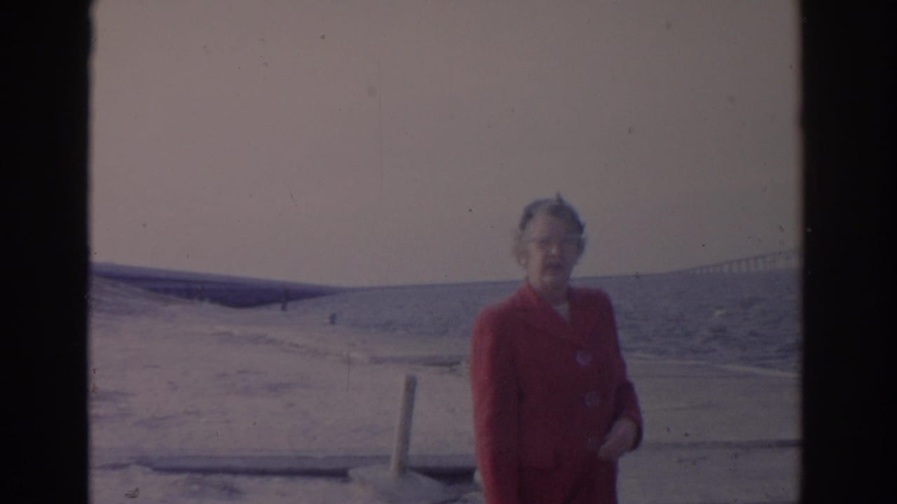 1962 Florida home-movie frame of an adult woman in a red coat near snowy shoreline, with water and a bridge in the background.