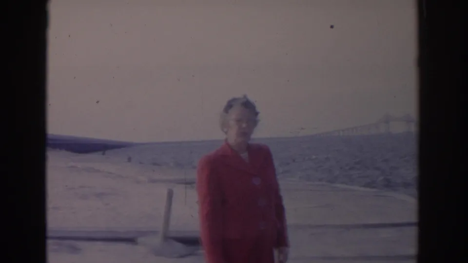In 1962 Florida home-movie footage, an older woman in a red coat faces the camera on a beach with choppy water and a distant bridge.