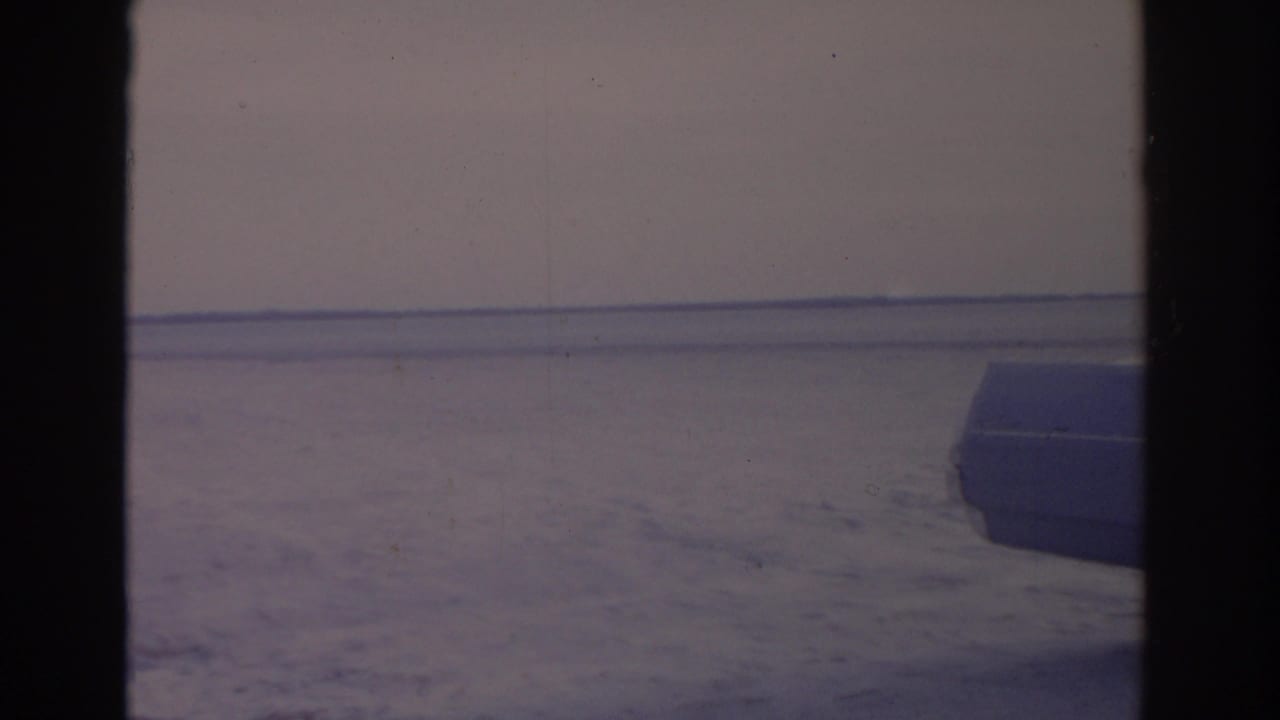 The camera leaves the woman and frames the sandy beach, flat waterline, pale sky, and part of a parked vintage car.