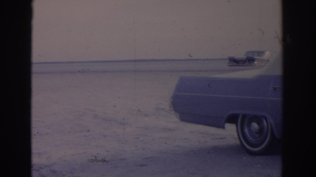 A waterfront view shows the rear of a parked car, open sand, distant water, and a small vehicle near the horizon in soft 8mm color.