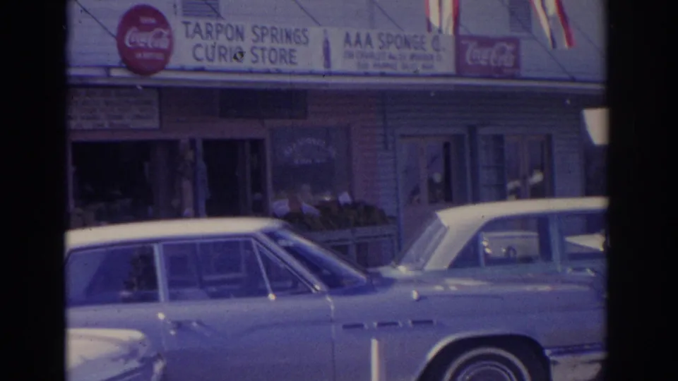 1962 Florida home-movie frame showing a light blue parked car, storefront signs for Tarpon Springs Curio Store and AAA Sponge Co., Coca-Cola logos, and small flags.