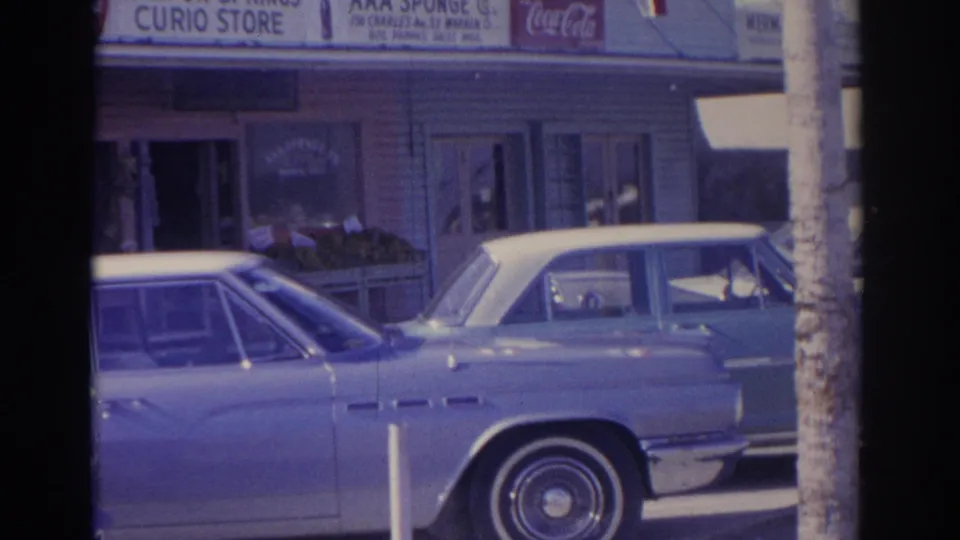 1962 Florida street frame with two parked early-1960s cars in front of shop windows, Coca-Cola signs, and Tarpon Springs retail signage.