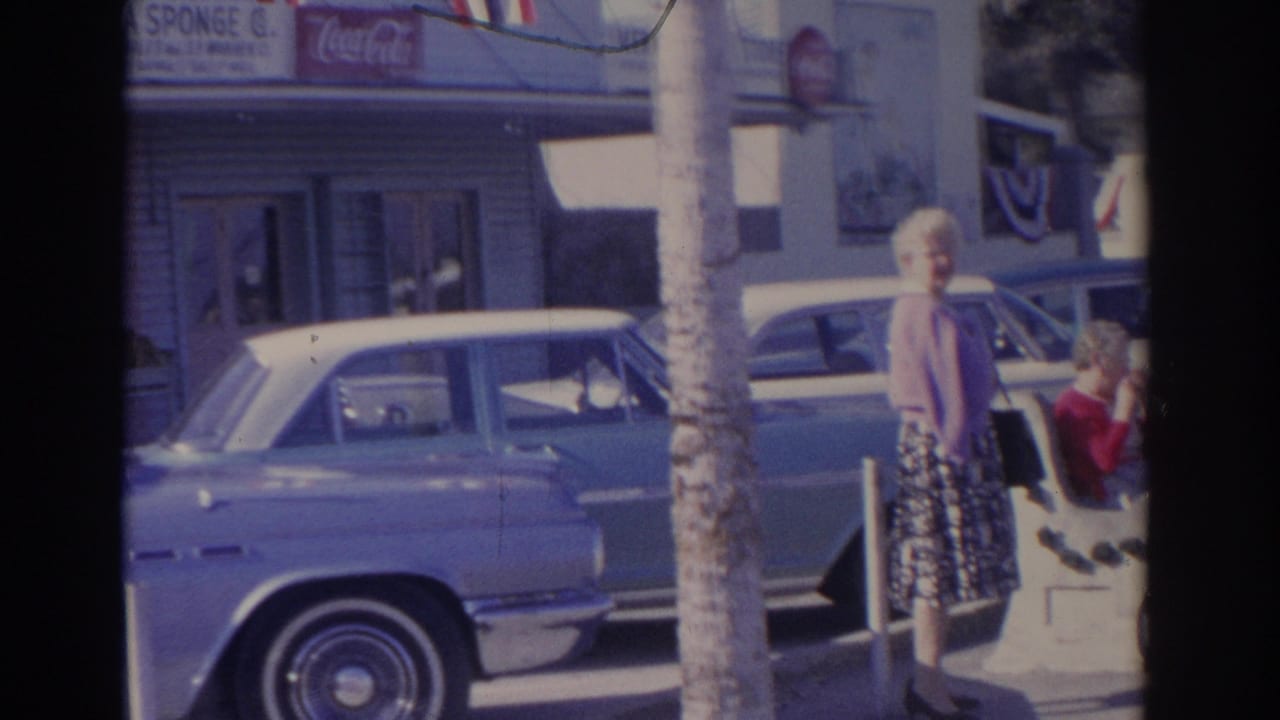 1962 Florida street frame with an older woman in a patterned skirt near parked cars, a palm trunk, storefront windows, patriotic bunting, and visible Coca-Cola signage.