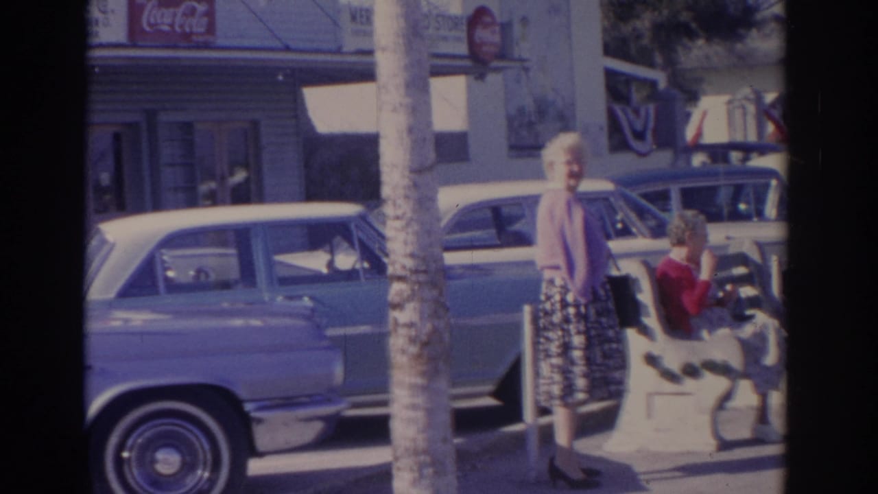 1962 Florida home-movie frame showing one woman standing and one seated on a bench near parked cars, a palm trunk, and a Coca-Cola sign.