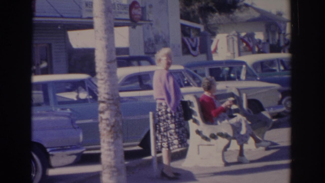 1962 Florida frame with a woman in a purple cardigan standing near a bench while another woman sits, with classic cars and storefront details behind.