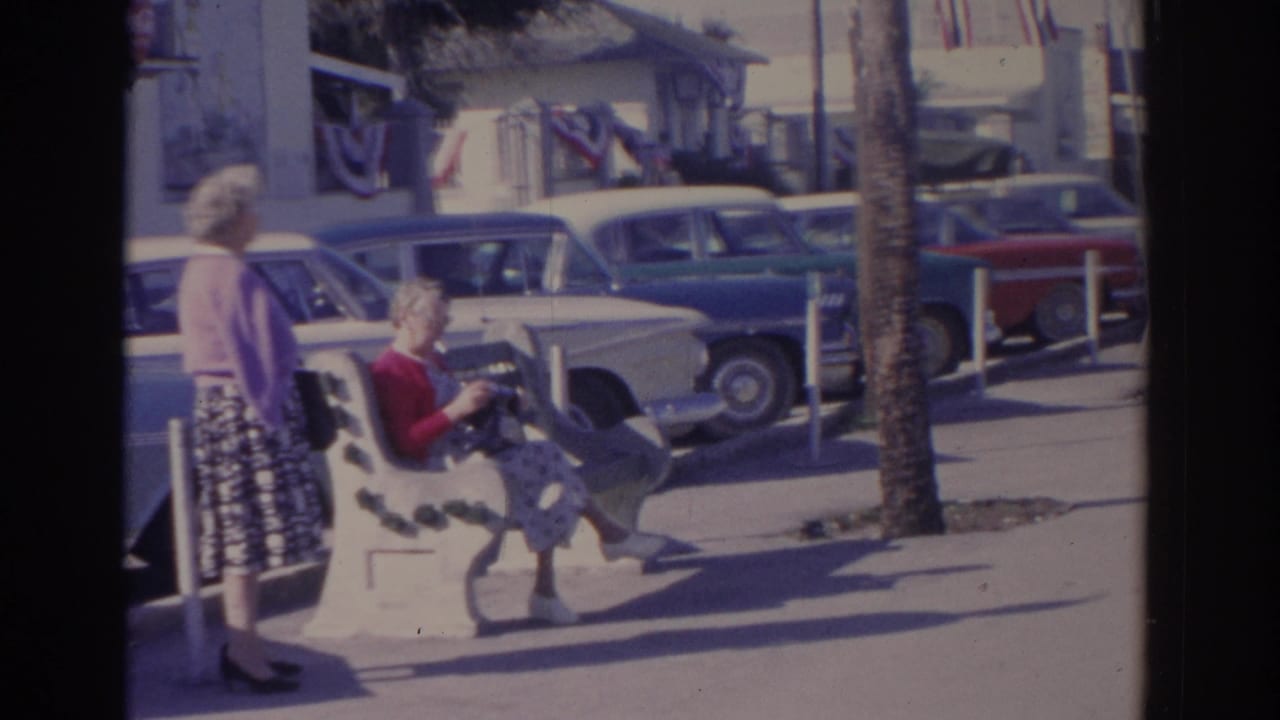 1962 Florida frame showing a seated woman and a standing woman on a sunny sidewalk, with classic cars, palm trunks, and storefront bunting in view.