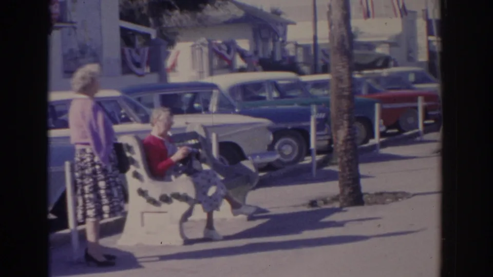 1962 Florida frame showing a seated woman and a standing woman on a sunny sidewalk, with classic cars, palm trunks, and storefront bunting in view.