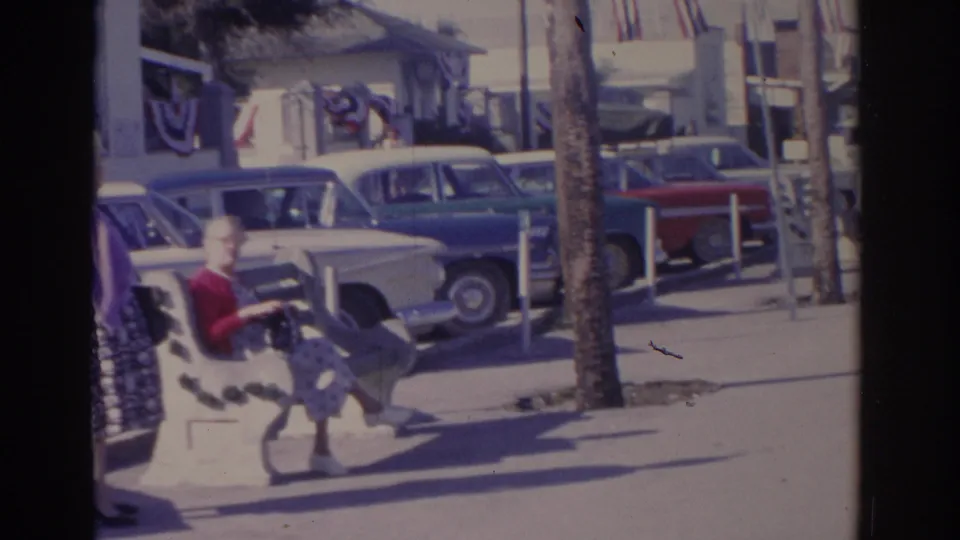 1962 Florida home-movie frame showing a woman on a patterned bench beside parked early-1960s cars and a palm-lined street.