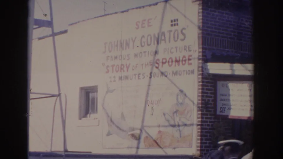 1962 Florida 8mm frame showing a large painted advertisement for Johnny Gonatos' Story of the Sponge on a white building wall.