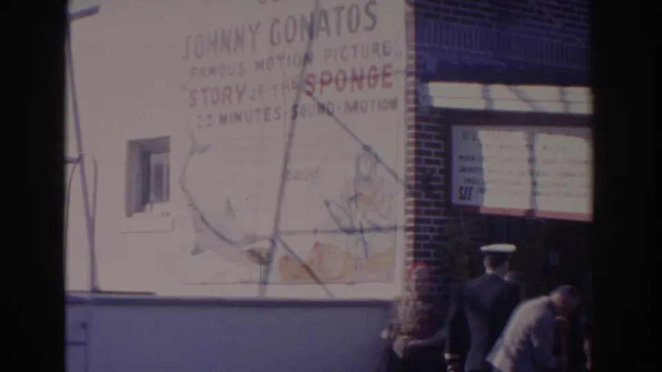 1962 Florida 8mm frame showing the lower part of a painted movie advertisement, a sailor, and people near a brick-front entrance.