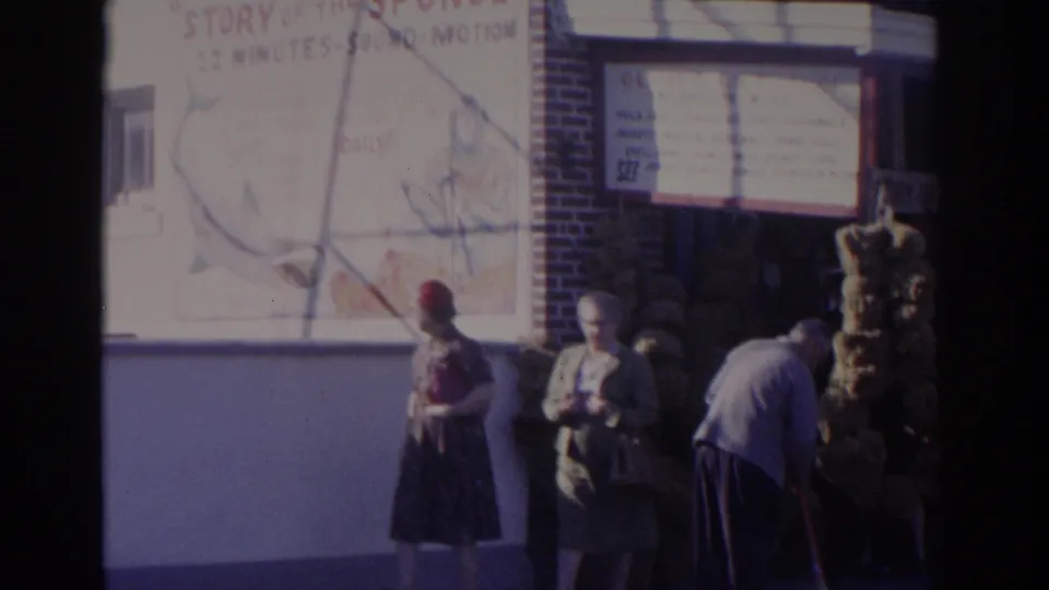 1962 Florida street scene with adults in period clothing below a painted Story of the Sponge wall advertisement on a commercial building.