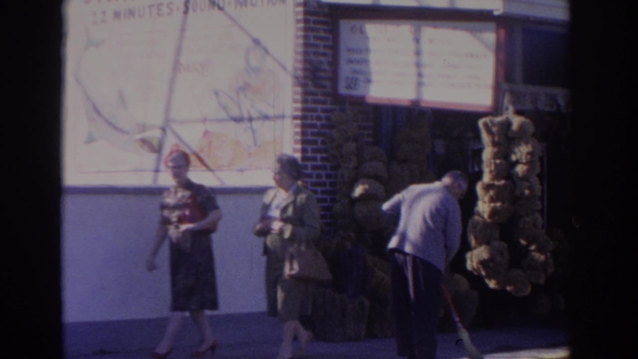 In 1962 Florida home-movie footage, two women in dresses pass a brick storefront with sponge displays as a man sweeps near the curb.