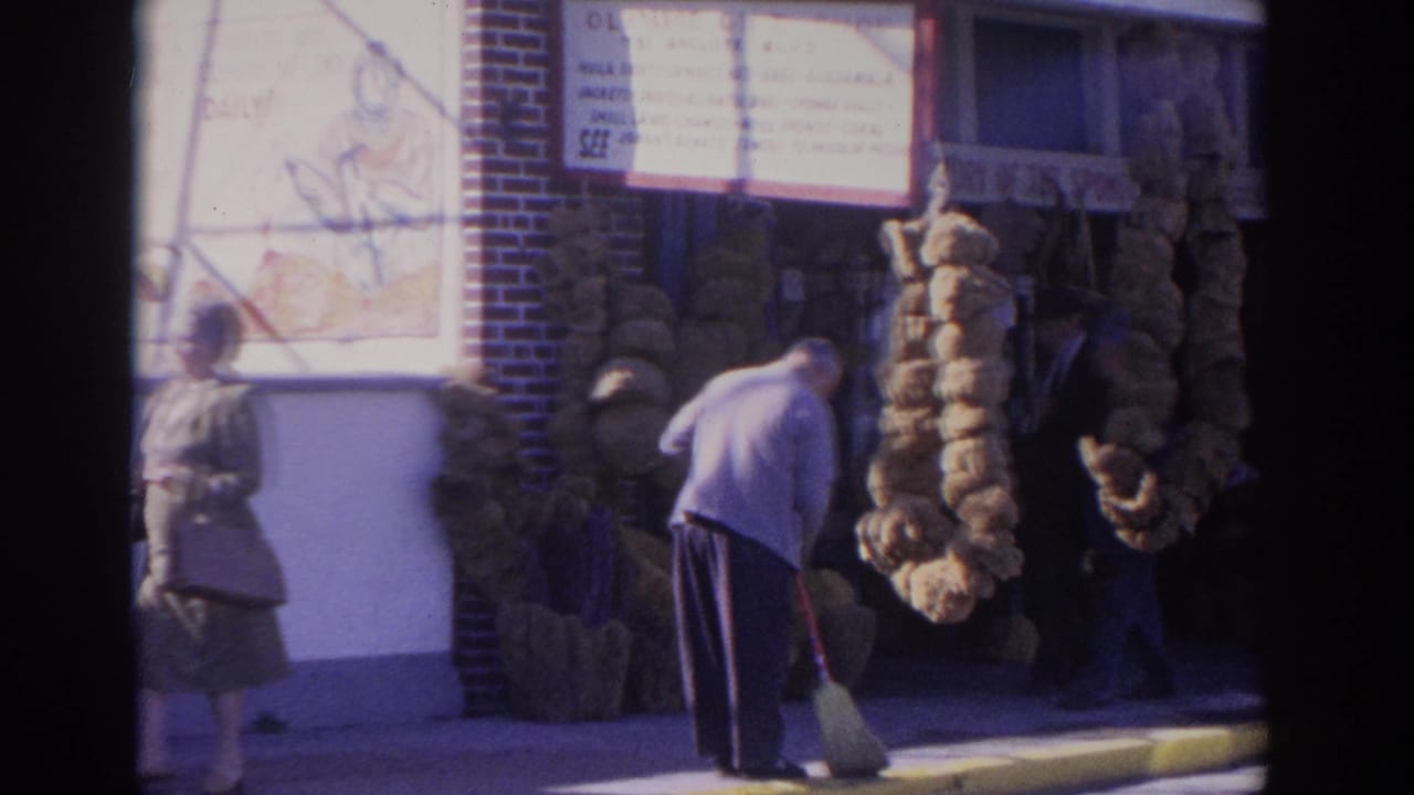 A man sweeps near stacked sponges outside a storefront as a woman walks by.