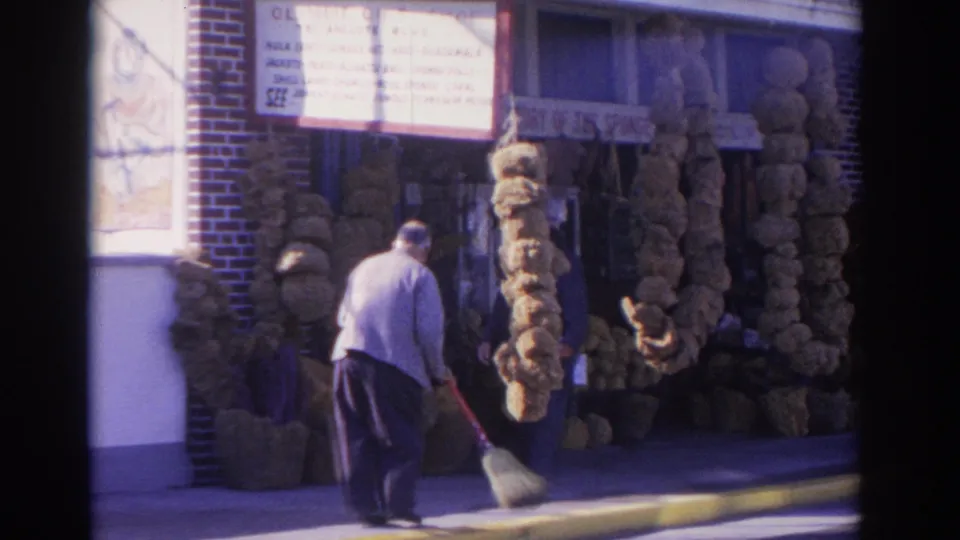 The home-movie frame shows a man sweeping along the curb in front of a Florida sponge shop display with large natural sponges around the entrance.