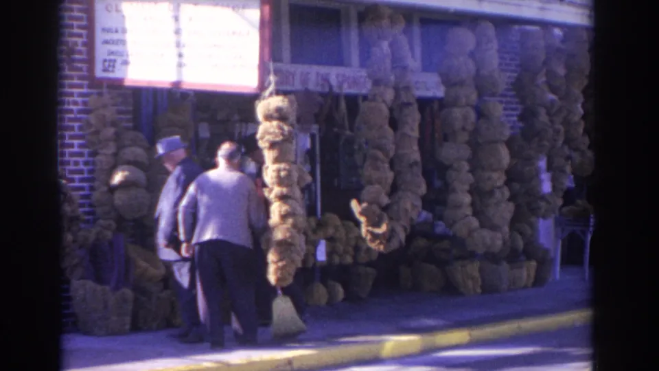 Near the end of the 1962 Florida clip, a man with a broom stands by the sponge shop entrance as another man in a hat faces him on the sidewalk.