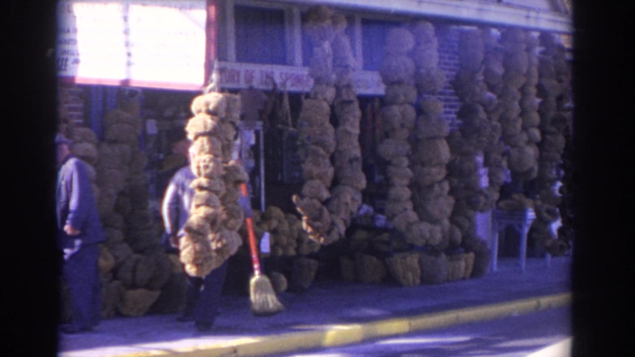 1962 Florida 8mm frame showing a man with a broom walking past a sponge shop display with a partial storefront sign.